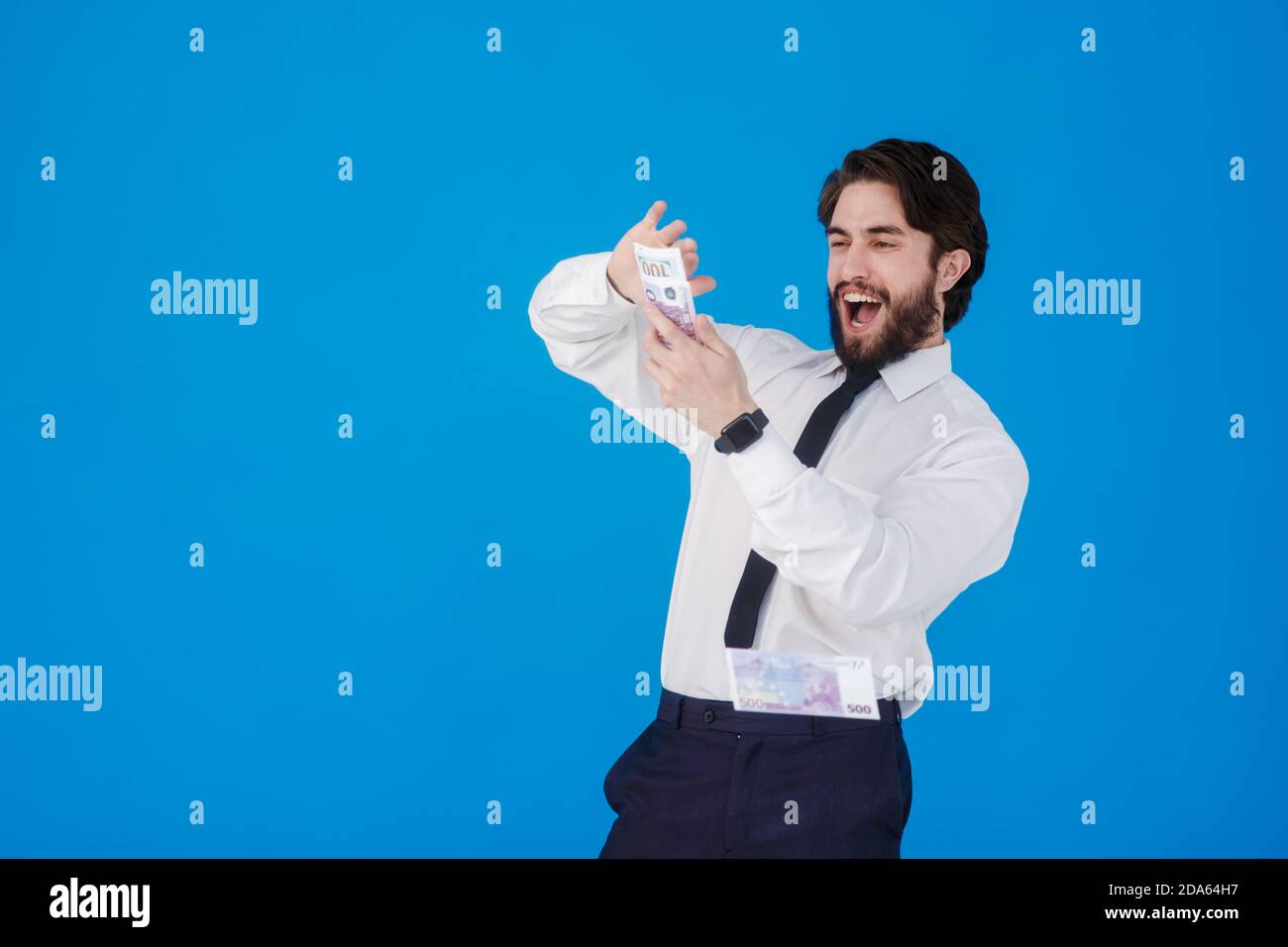 Ein junger fröhlicher bärtiger Geschäftsmann in einem weißen Hemd und schwarzer Krawatte auf blauem Hintergrund spritzt Geld. Verrückter Kerl wirft Geldscheine. Studio Stockfoto