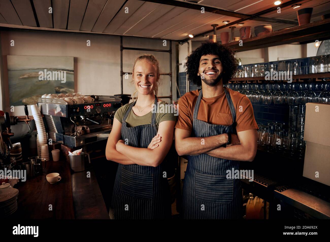 Portrait von jungen weiblichen und männlichen Mitarbeitern mit Schürze stehend Hinter dem Tresen im Café mit gekreuzten Händen und Blick auf die Kamera Stockfoto