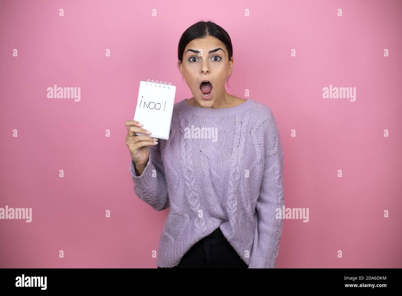 Schöne Frau trägt einen lässigen violetten Pullover auf rosa Hintergrund Erschrocken und überrascht halten Notebook ohne Wort Nachricht Stockfoto