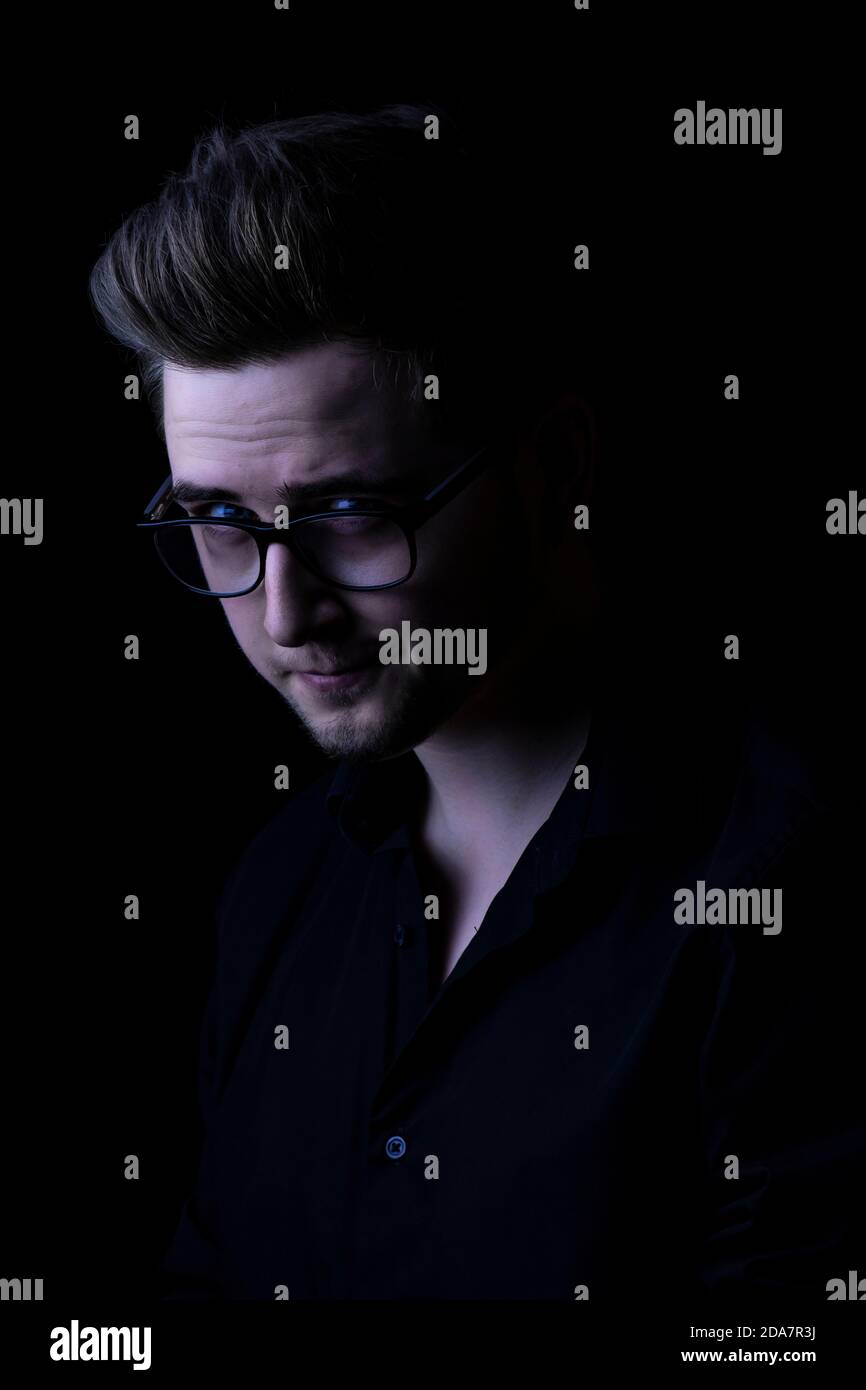 Mann starrte die Kamera schräg an und trug ein schwarzes Hemd vor einem schwarzen Hintergrund Stockfoto
