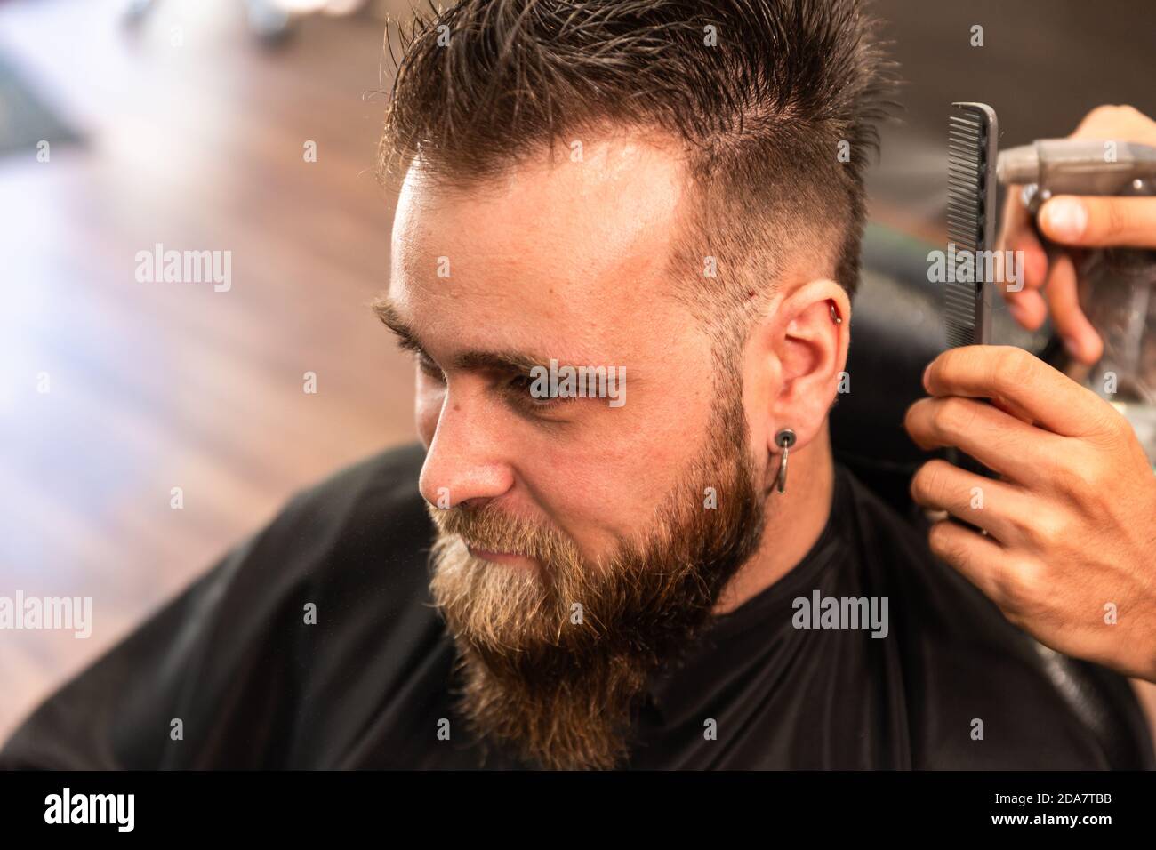 Kaukasischer Mann mit Bart wird in einem Friseurladen gekämmt Und mit einem Wasserspender von den Händen gesprüht Ein ethnischer hispanischer Barbier.der Kunde trägt ein Ohr Stockfoto