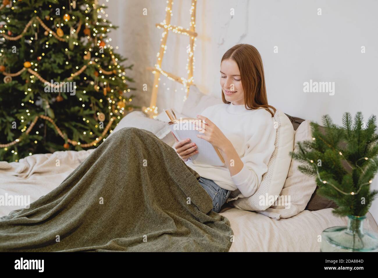 Attraktive junge Frau liegt im Bett mit karierten Beinen und liest am Weihnachtsmorgen ein Buch und liest ein Buch. Stockfoto