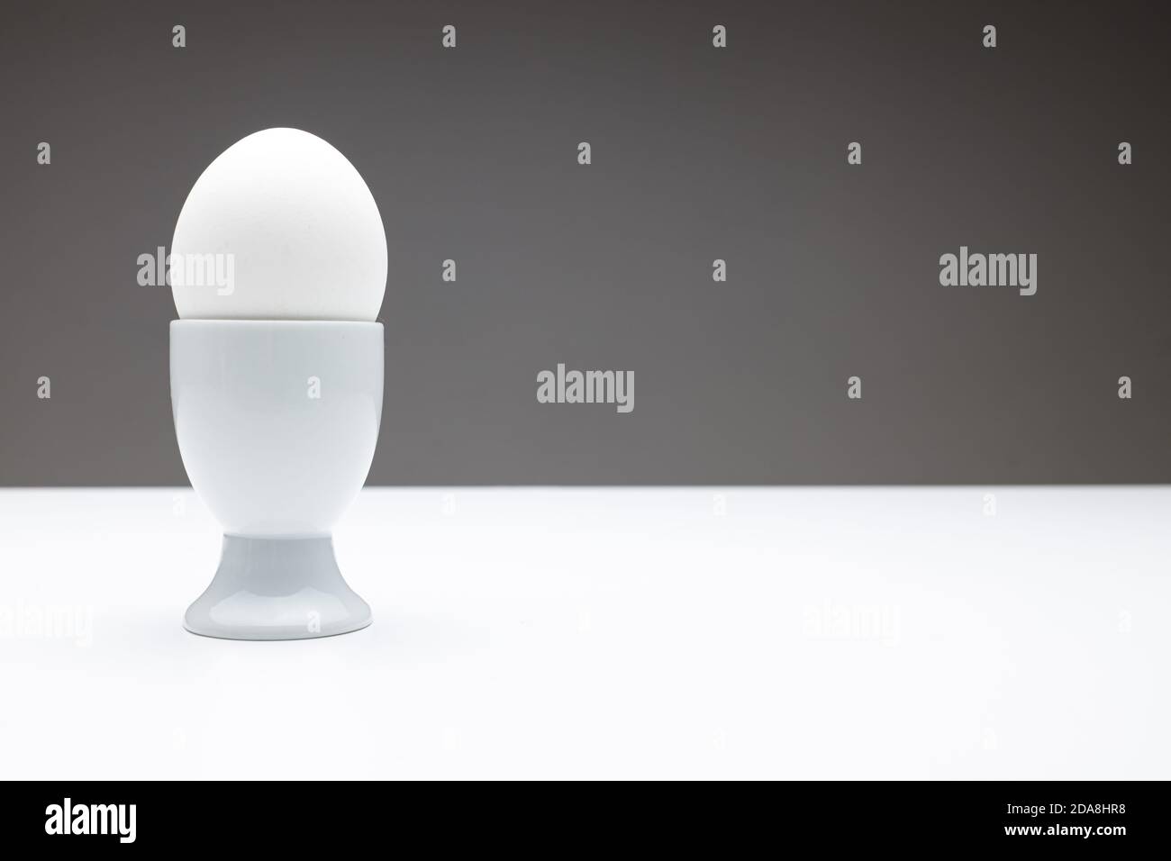 Vorderansicht eines Eierbechers mit einem weiß gekochten Ei auf einem dunkelgrauen Hintergrund auf einem weißen Tisch mit weichem Licht, als Vorlage für die Bemalung von Gesichtern oder Emoti Stockfoto