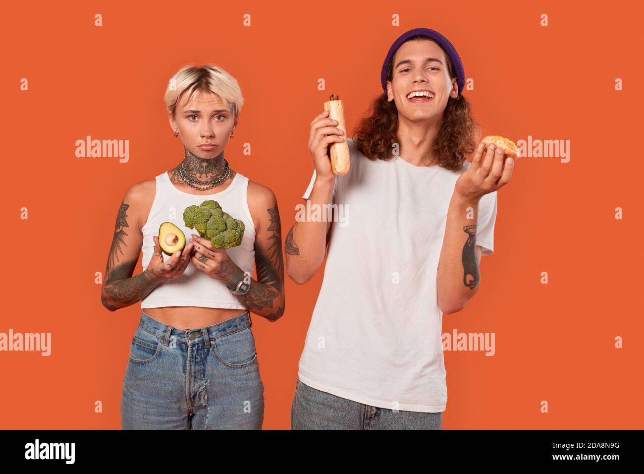 Freistil. Junge tätowierte Paar stehend isoliert auf orange Freundin hält Gemüse gesunden Lebensstil vs Freund hält Hot Dog und Donut Stockfoto