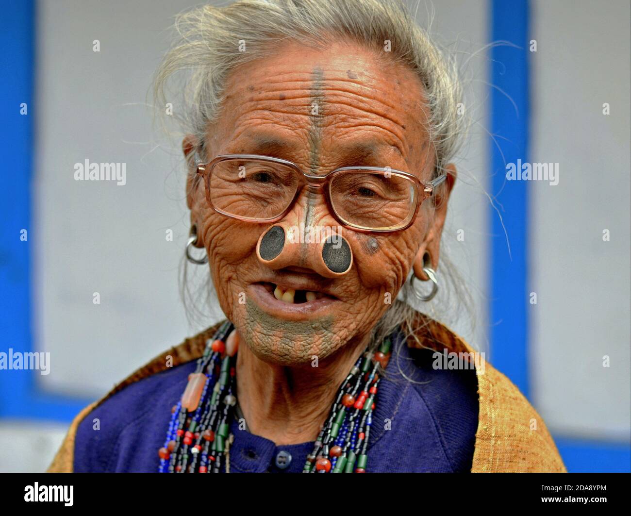 Alte nordöstliche indische Apatani Stammesfrau mit schwarzen hölzernen Nasenstopfen und traditionellem Gesichts-Tattoo trägt moderne Brillen und lächelt für die Kamera. Stockfoto