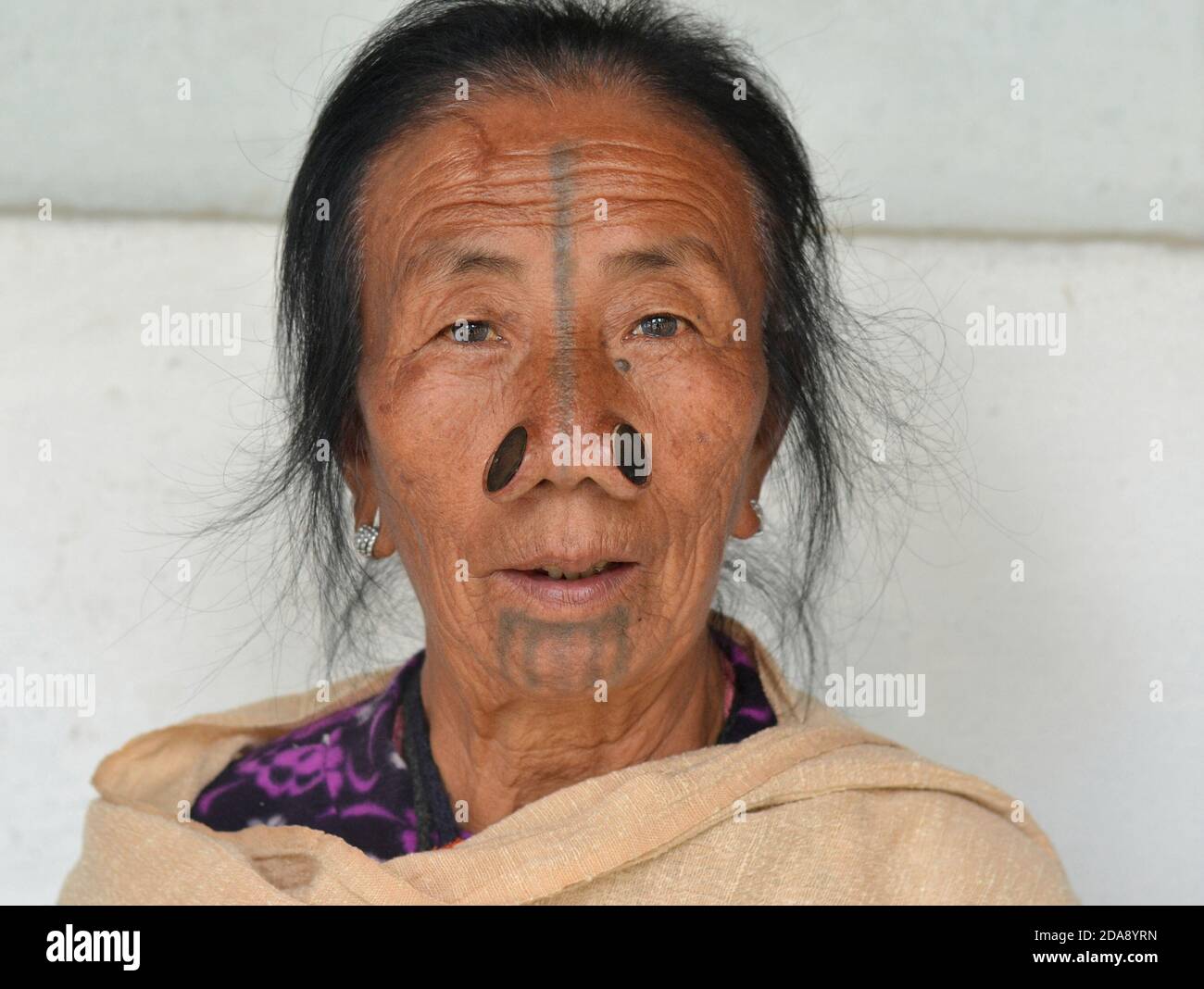 Ältere nordöstliche indische Apatani ethnische Minderheit Stammesfrau mit schwarzen hölzernen Nasenstopfen und traditionellen Gesicht Tattoo Posen für die Kamera. Stockfoto