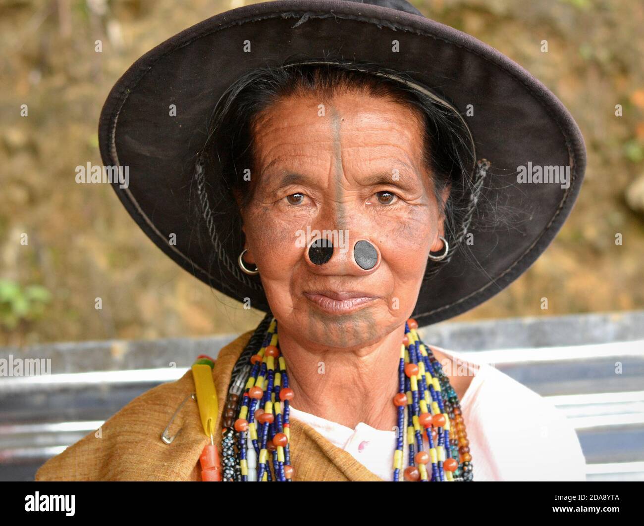 Ältere nordöstliche indische Apatani-Stammesfrau mit schwarzen Nasenstopfen und traditionellen Gesichts-Tattoos trägt einen modernen Sonnenhut und Posen für die Kamera. Stockfoto