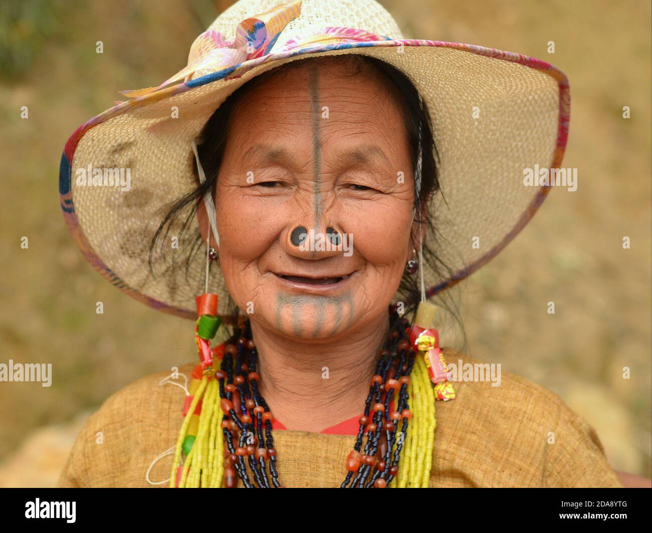 Ältere nordöstliche indische Apatani-Stammesfrau mit schwarzen Nasenstopfen und traditionellen Gesichts-Tattoos trägt einen modernen Sonnenhut und lächelt für die Kamera. Stockfoto