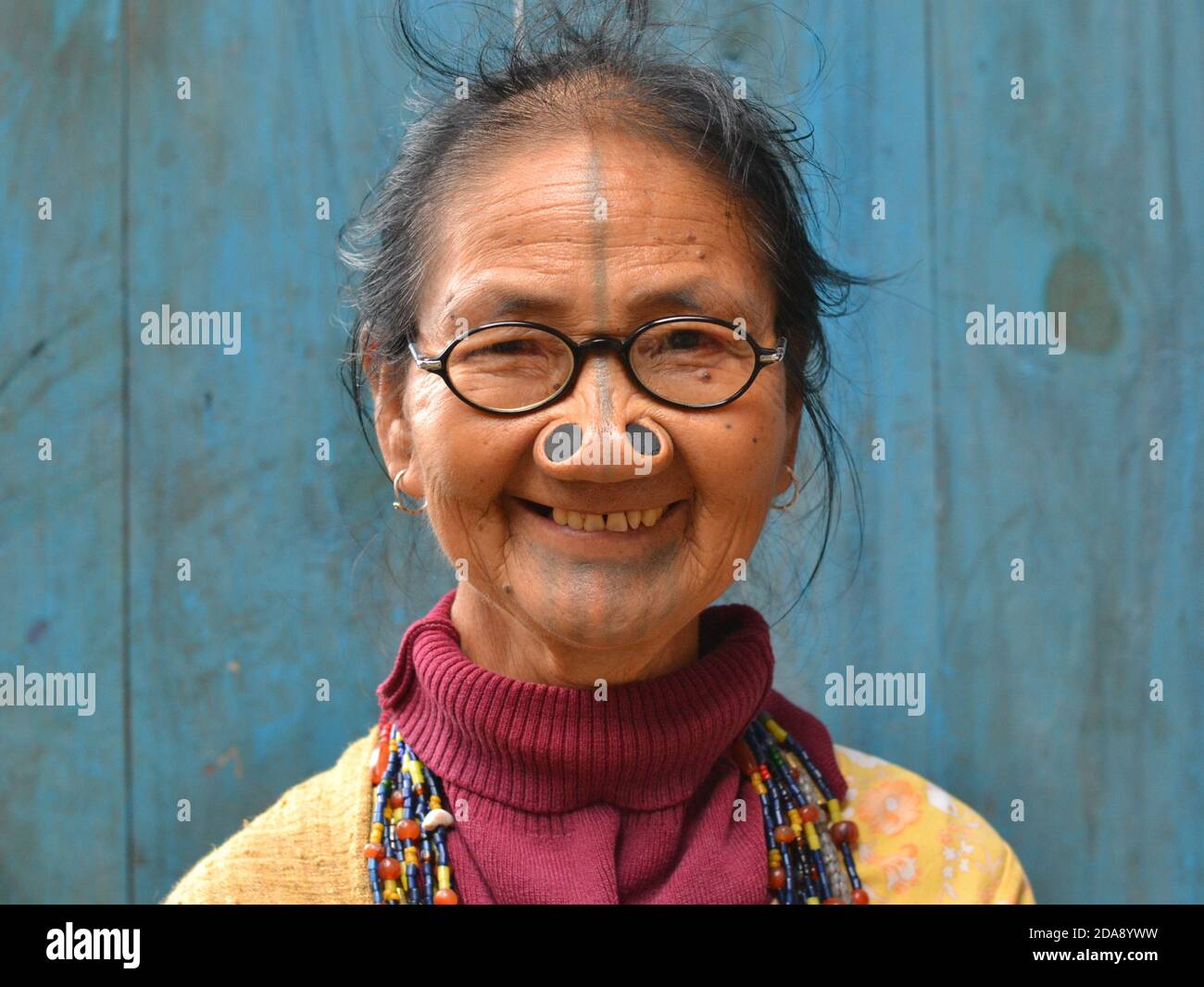 Alte nordöstliche indische Apatani Stammesfrau mit schwarzen hölzernen Nasenstopfen und traditionellem Gesichts-Tattoo trägt moderne Brillen und lächelt für die Kamera. Stockfoto