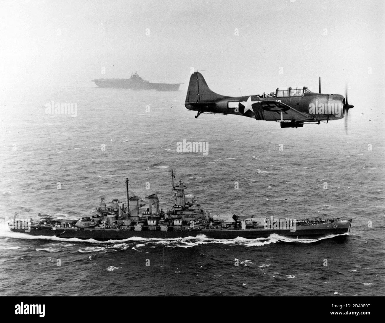 Eine US Navy Douglas SBD-5 Dauntless von Bombengeschwader VB-16 fliegt eine Anti-U-Boot-Patrouille tief über dem Schlachtschiff USS Washington (BB-56) auf dem Weg zur Invasion der Gilbert-Inseln, 12. November 1943. Das Schiff im Hintergrund ist die USS Lexington (CV-16), der Heimatträger des Flugzeugs. Notieren Sie sich die Tiefenladung unter dem SBD. 12. November 1943 Stockfoto