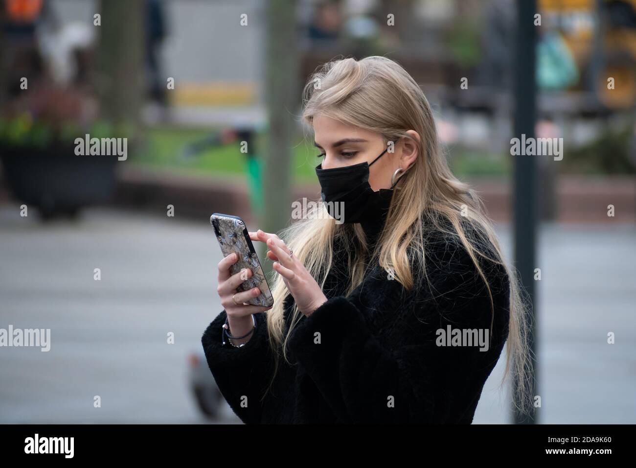 Schöne junge blonde Mädchen mit Maske Überprüfung der App Stop Covid auf dem Smartphone auf der Straße während Covid oder Coronavirus Notfall, Verfolgung App Stockfoto