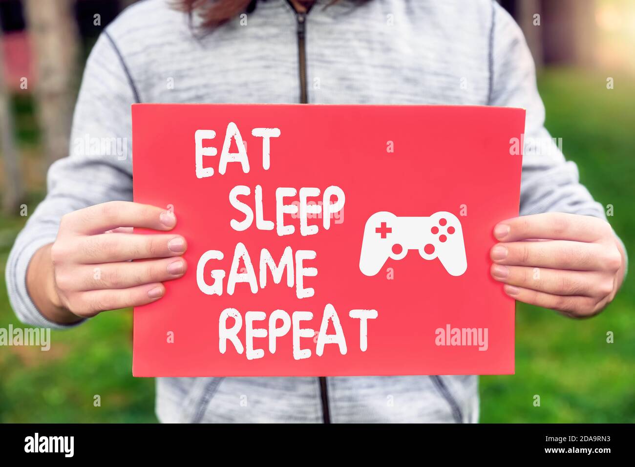 Männliche Teenager Gamer hält ein Banner mit essen, schlafen, Spiel, wiederholen Zitat Nachricht mit einem Gamepad-Symbol. Spielabhängigkeit oder Abhängigkeitskonzept. Stockfoto