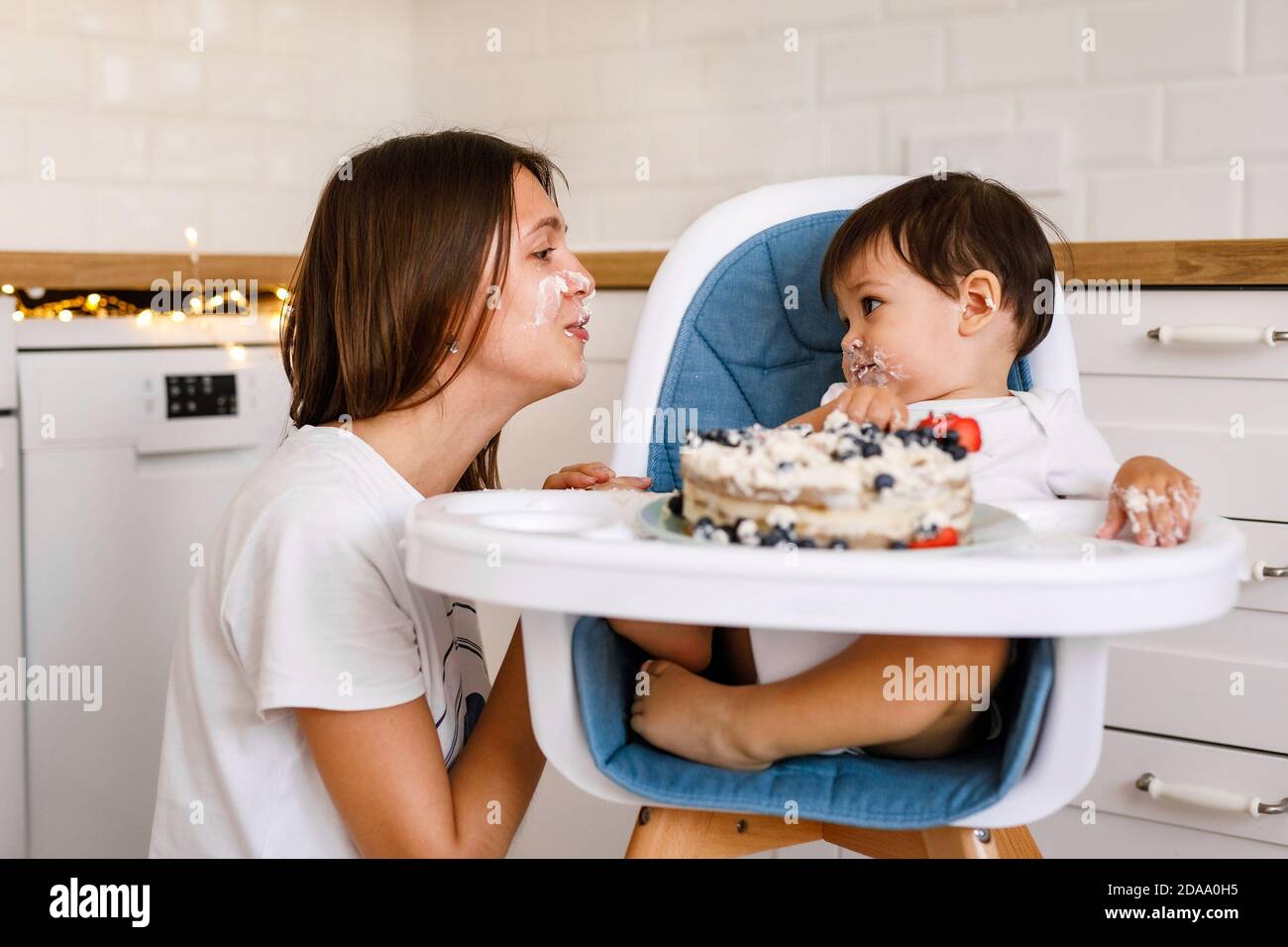 Glückliche Mutter mit einem Jahr alten Baby feiert mit ersten Geburtstag mit weißer Küche auf Hintergrund. Stockfoto