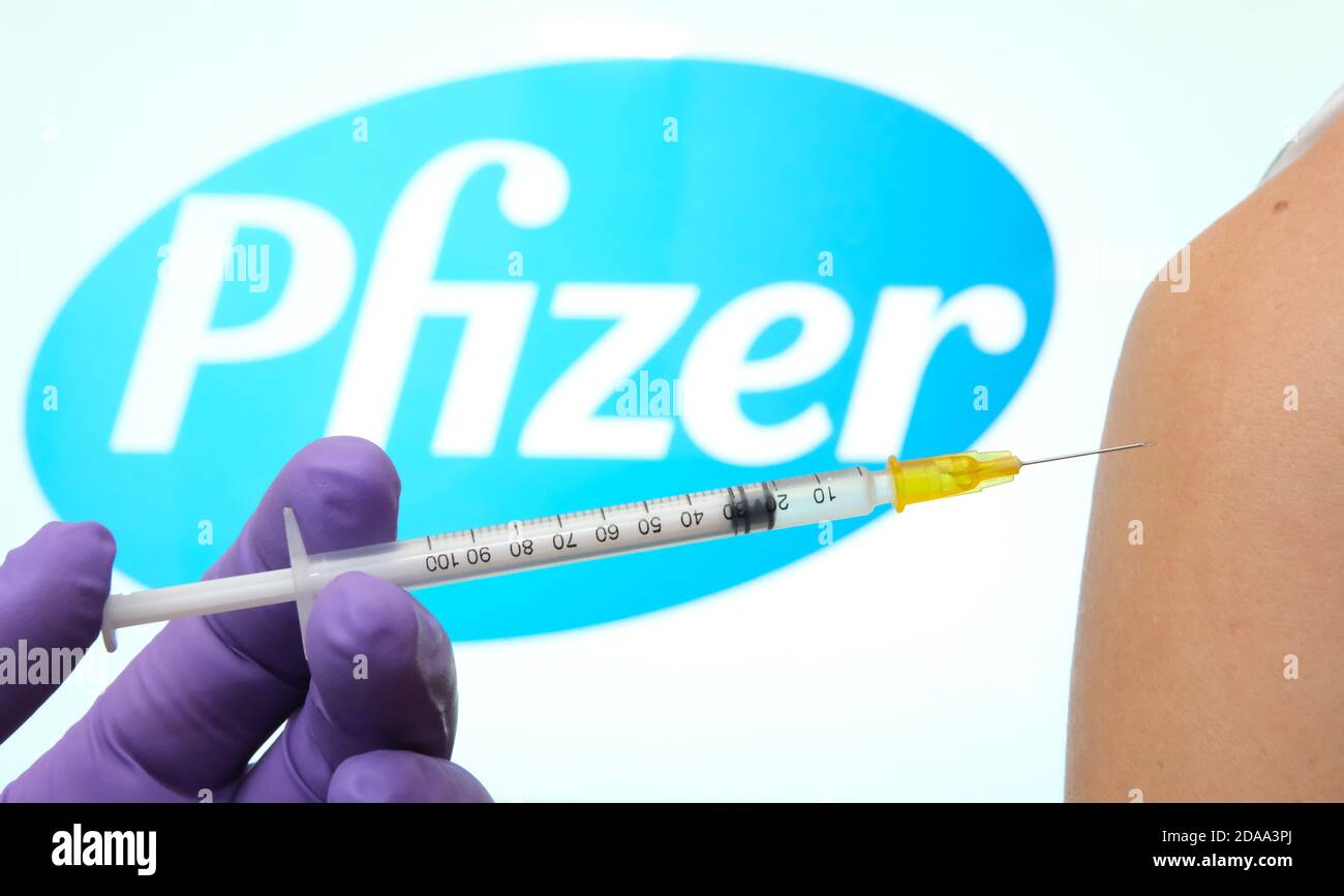 Pfizer BioNTech Covid-19 Impfstoffkonzept. Hand hält eine Spritze gegen die Frau Schulter, mit verschwommenem Pfizer-Logo auf der Rückseite. Stockfoto