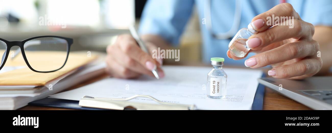 Arzt schreibt Dokument an Bord am Tisch und halten Flasche mit Impfstoff gegen Coronavirus-Infektion Nahaufnahme Stockfoto