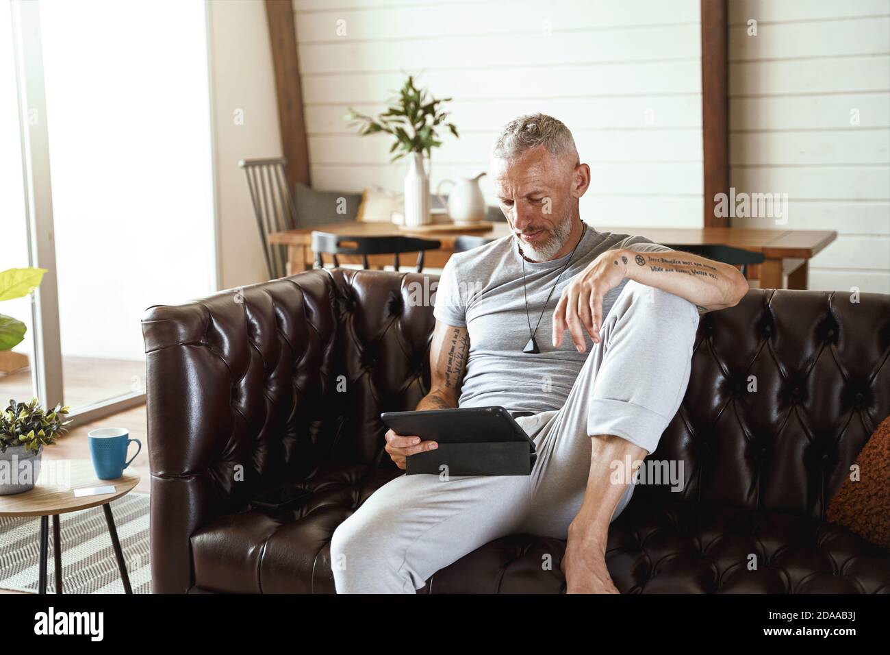 Fokussierter Kaukasischer Mann mittleren Alters in legerer Kleidung mit digitalen Tablet, während Sie auf dem Sofa im Wohnzimmer von sitzen Seine moderne Wohnung Stockfoto