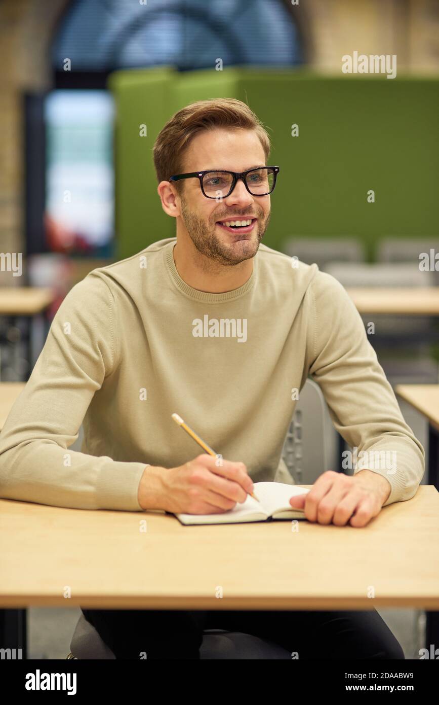Business Education Konzept. Fröhlicher männlicher Büroangestellter sitzt am Schreibtisch im Coworking Space, macht Notizen und lächelt während des Corporate Trainings oder Seminars Stockfoto