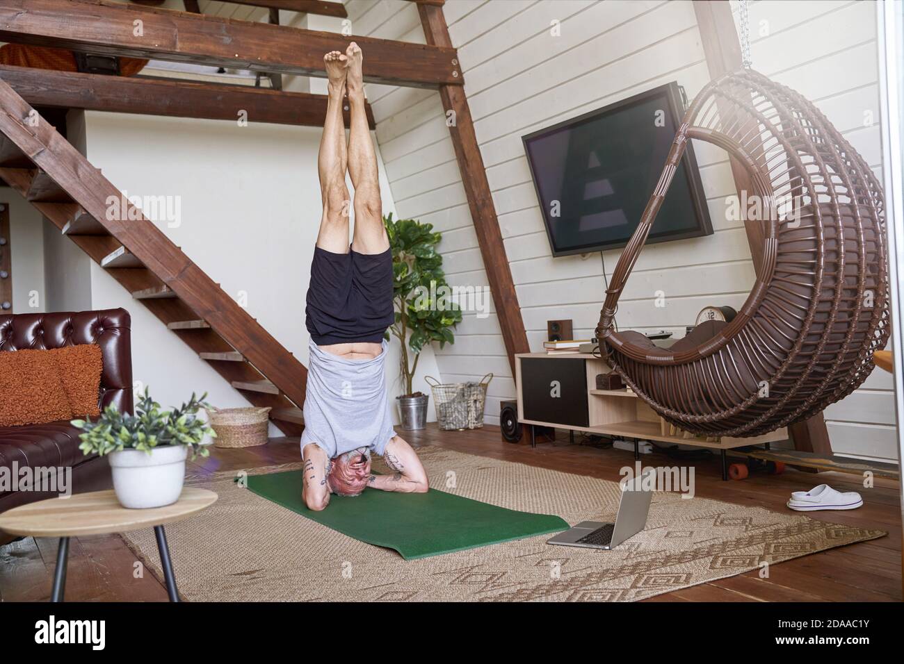 In voller Länge eines sportlichen Mann mittleren Alters in Kopfstand Pose stehen, während Online-Video-Tutorial auf Laptop, die Ausübung im Wohnzimmer Stockfoto