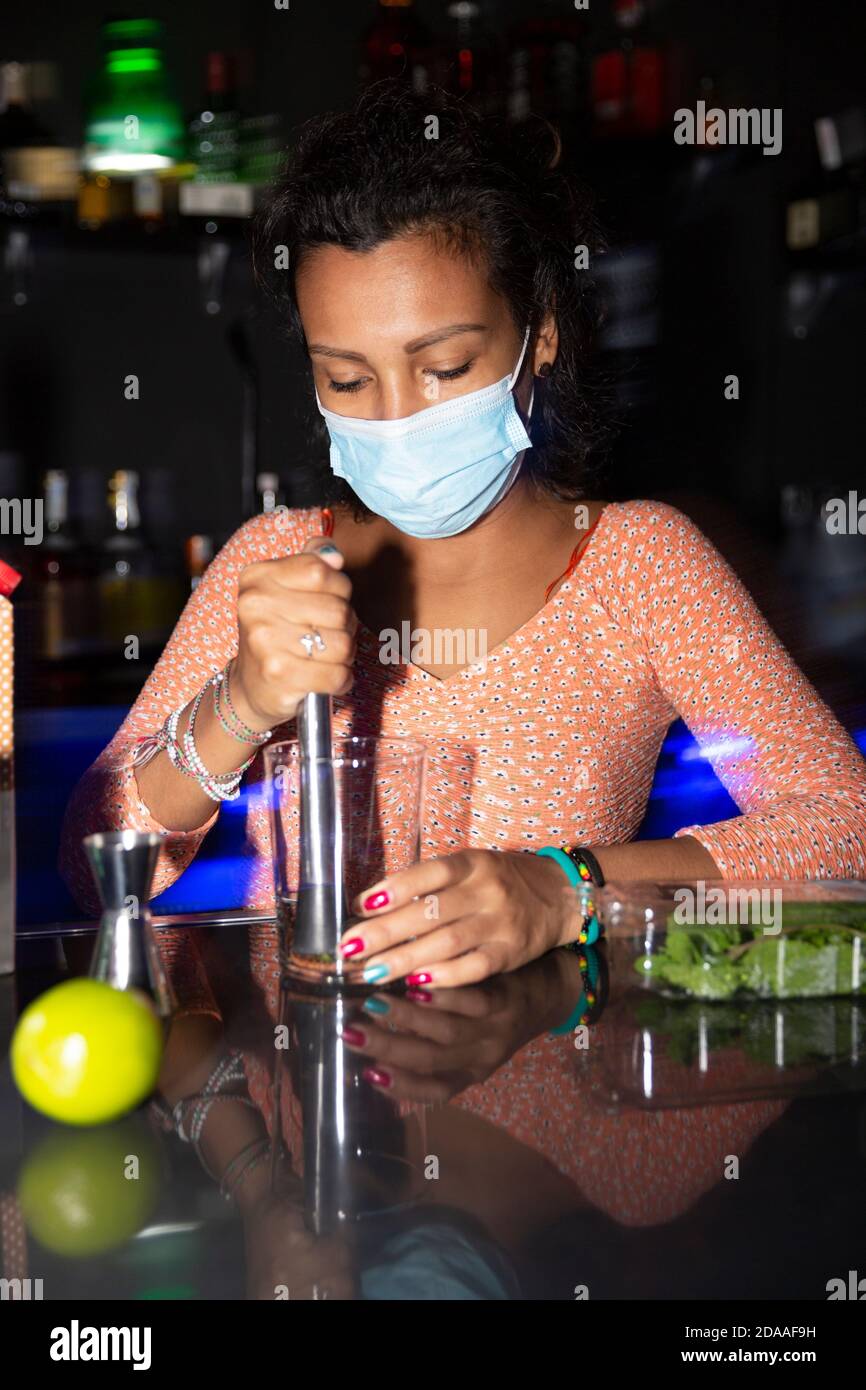 Porträt einer jungen lateinamerikanischen Kellnerin, die in einer Disco-Bar einen Mojito vorbereitet. Sie trägt eine medizinische Maske. Stockfoto