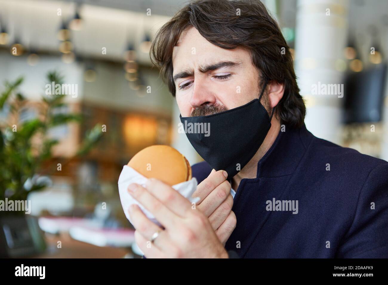 Mann mit einer Gesichtsmaske auf dem Kinn wegen Covid-19 Essen eines Burger im Restaurant Stockfoto