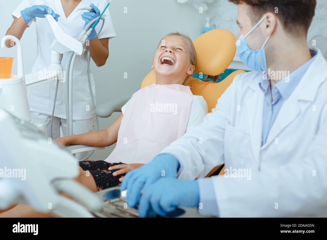 Junge Ärztin und Krankenschwester in Schutzmasken, Handschuhen und Uniformen schaut auf lachendes Mädchen Stockfoto