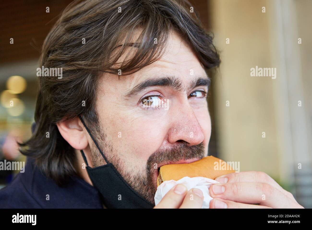 Hungriger Mann essen Burger mit Maske auf seinem Kinn wegen Zur Pandemie Covid-19 Stockfoto