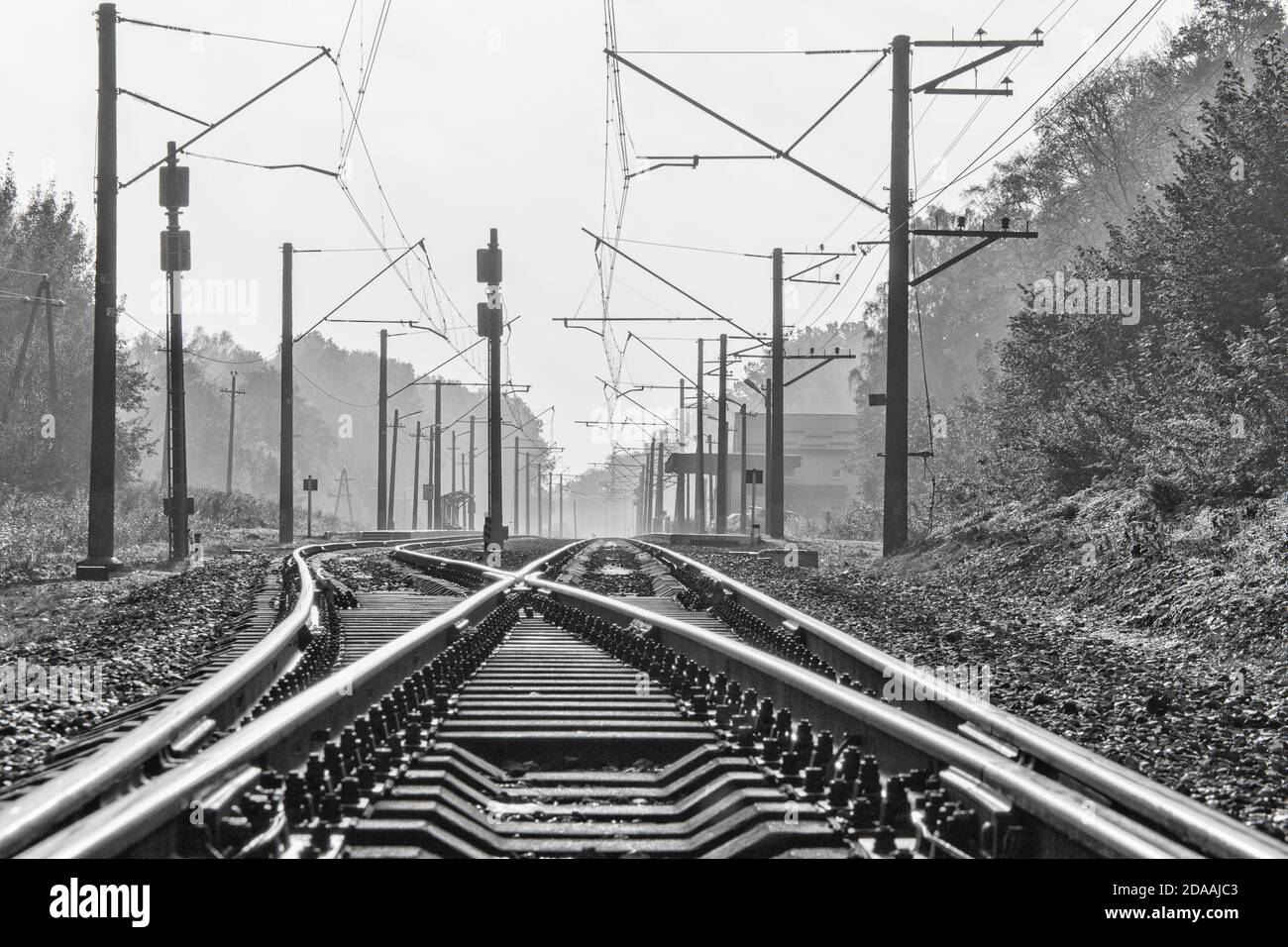 Zwei Bahngleise mit Strommasten und Kabeln in der Nähe des Bahnhofs eines kleinen Townships. Schwarzweiß-Foto Stockfoto