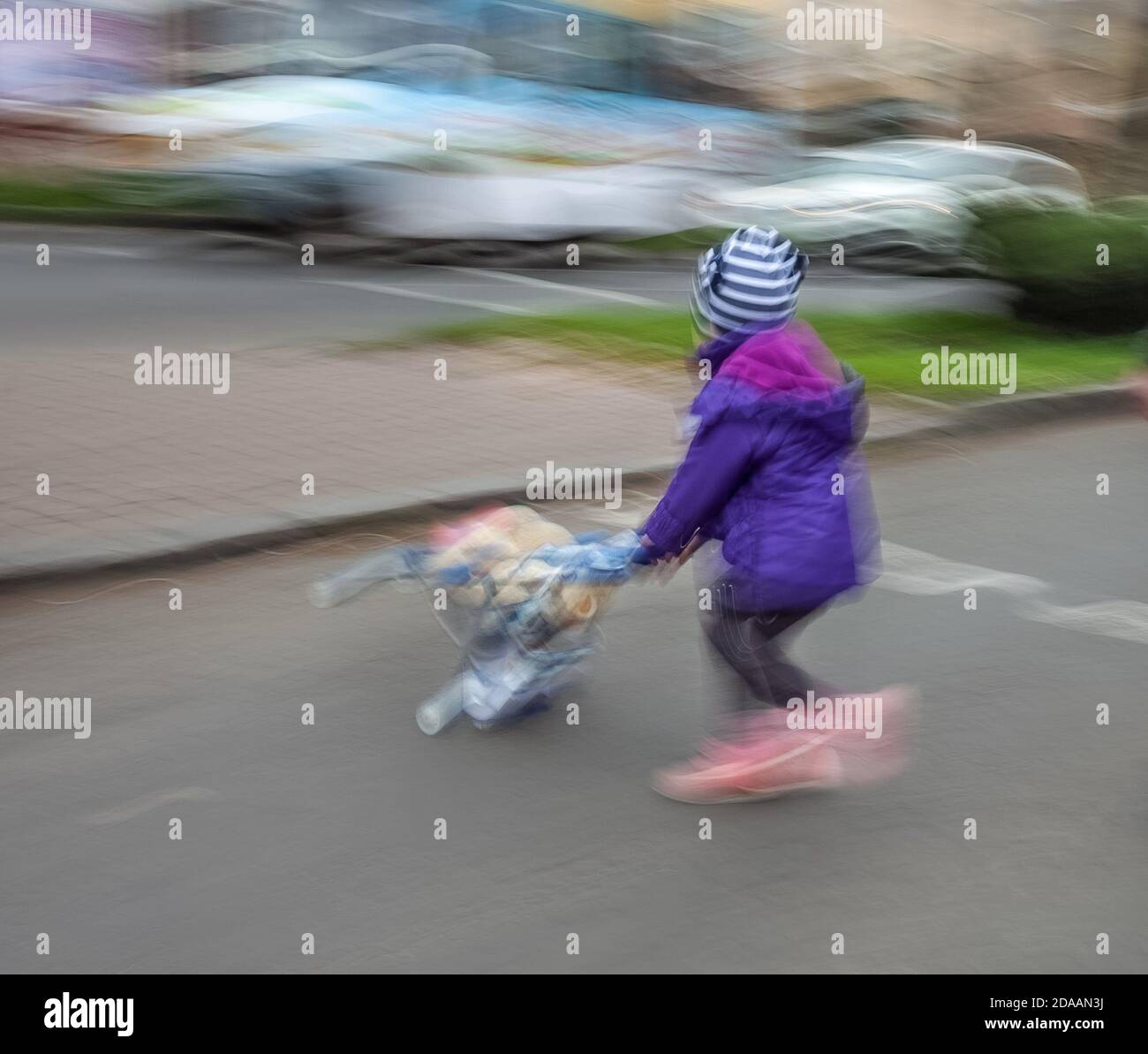 Kleines Mädchen mit Spielzeug Kinderwagen über die Straße. Absichtliche Bewegungsunschärfe Stockfoto