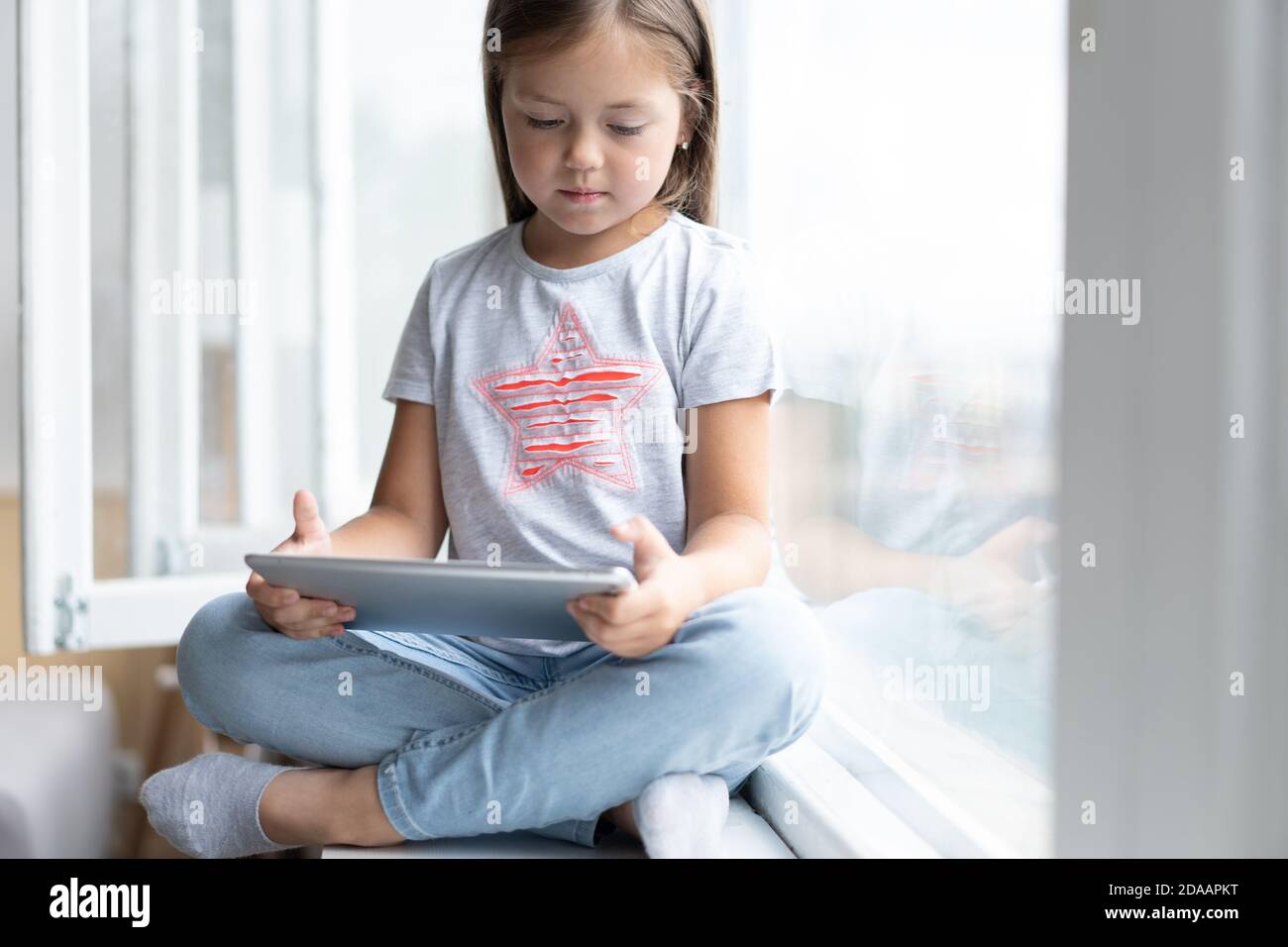 Nettes kleines Kind mit einem digitalen Tablet in einem Zuhause mit großem Fenster. Stockfoto