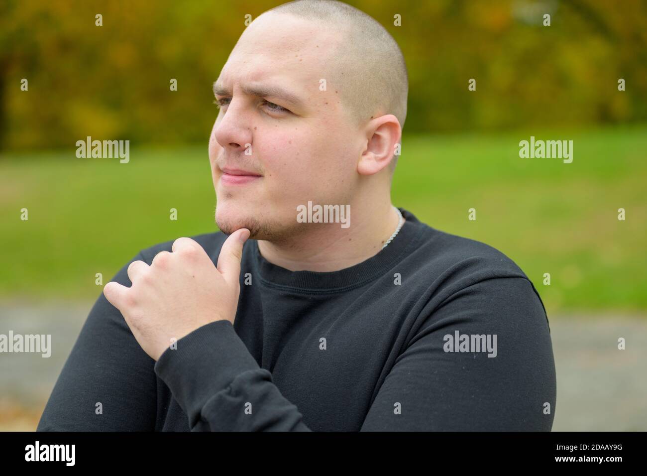Nachdenklicher junger Mann, der mit seinem auf die Seite schaut Hand an sein Kinn und ein kontemplativer Ausdruck im Freien in Ein Garten oder Park Stockfoto