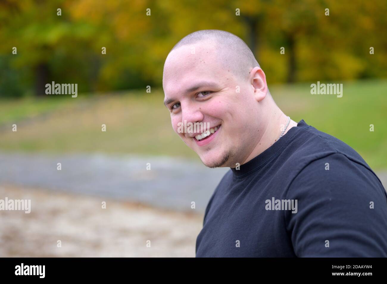 Lächelnd freundlich verdickt junger Mann mit einem rasierten Kopf suchen Seitlich an der Kamera im Freien in einem Park im Herbst Stockfoto