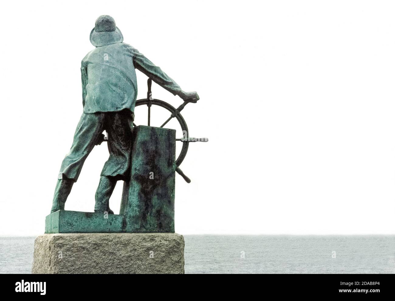 Diese Bronzestatue eines Fischers am Schiffsrad blickt auf den Hafen in Gloucester, Massachusetts, USA, als Denkmal für die Tausenden von Fischern, die während der ersten drei Jahrhunderte (1623-1923) der Geschichte dieser Stadt in Neuengland auf See verloren gegangen sind. Aus einem biblischen Psalm stammt die bekannte Inschrift des Denkmals: "Die in Schiffen ins Meer hinunterfahren." Es ist auf einem großen Granitblock gemeißelt, in dem die 2.4 Meter hohe Statue des englischen Bildhauers Leonard Craske steht. Dieses berühmte Denkmal steht im US National Register of Historic Places. Stockfoto
