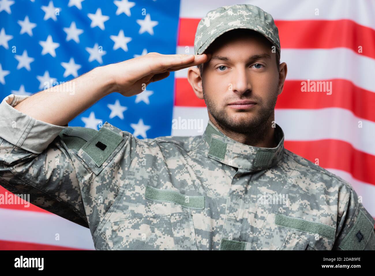 Patriotischer Militärmann in Uniform und Mütze, der in der Nähe grüßt amerikanische Flagge auf verschwommenem Hintergrund Stockfoto