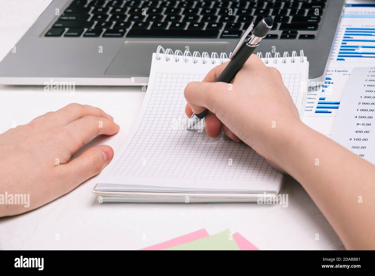 Die Frau am Tisch schreibt in einem Notizbuch. Mann, der einen Stift über ein leeres Notizbuch hält. Schreibtisch mit Computer und Finanzkarten Stockfoto