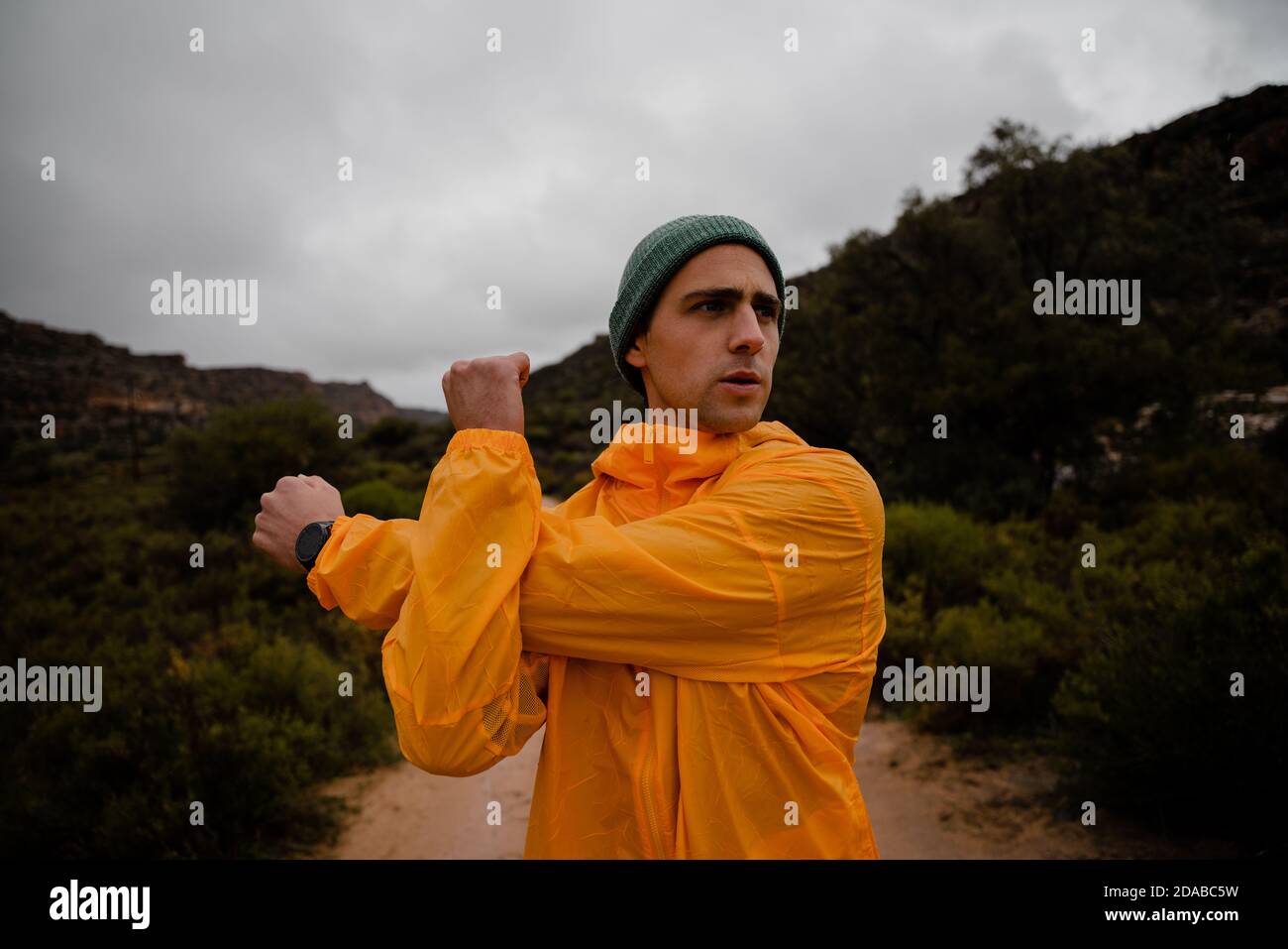 Fokussierter junger männlicher Wanderer, der nach einer steilen Wanderung Schultermuskel dehnt Bergpfad bei bewölktem Wetter mit üppigen Büschen Stockfoto