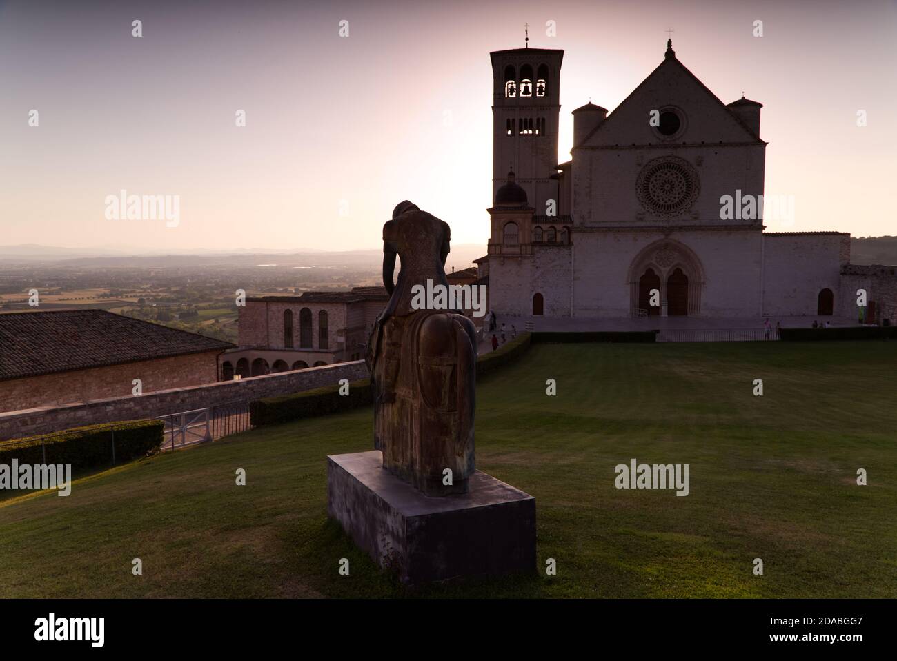 Die Statue des heiligen Franziskus mit der Basilika von Assisi im Hintergrund bei Sonnenuntergang Stockfoto