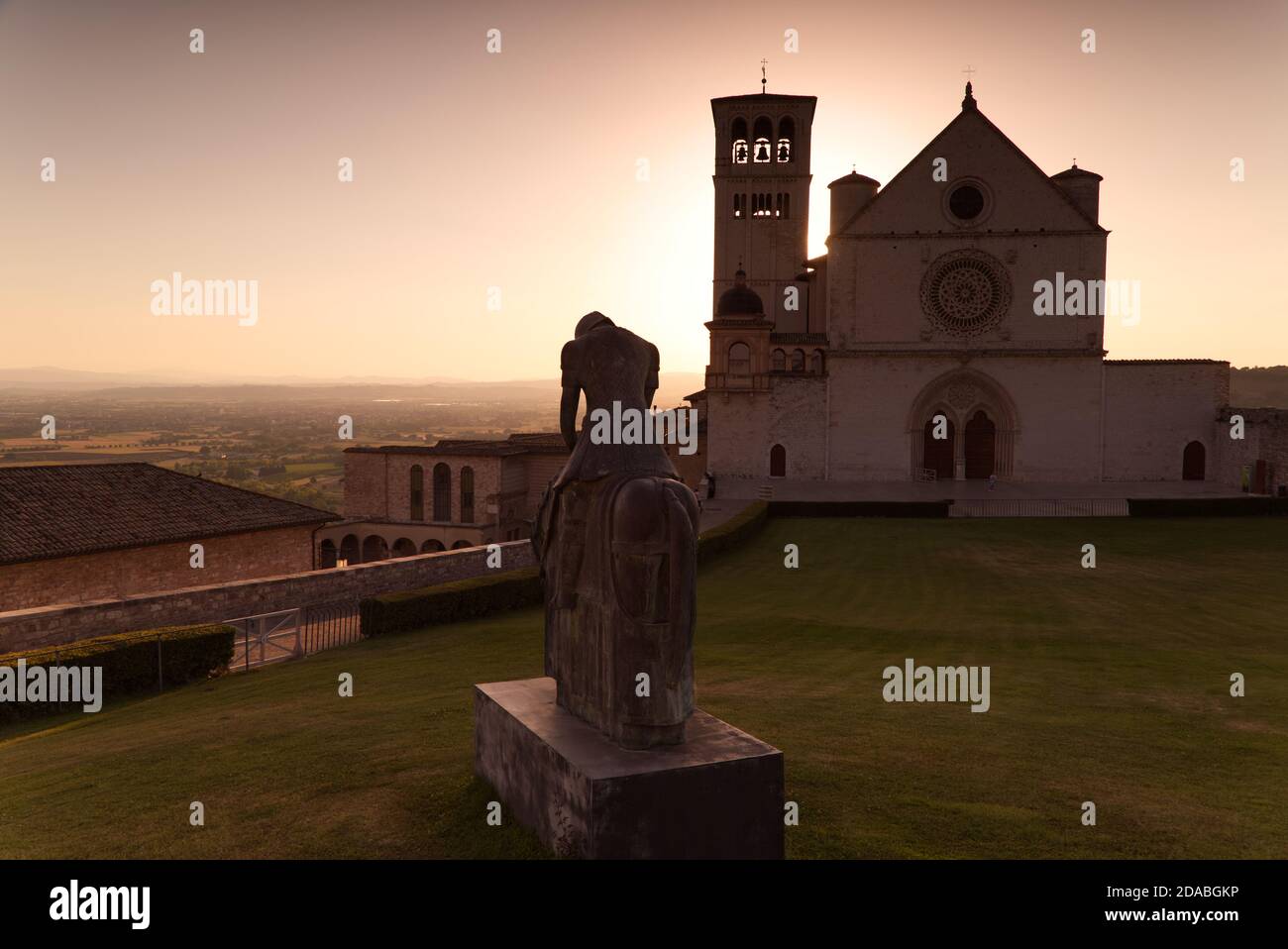 Die Statue des heiligen Franziskus mit der Basilika von Assisi im Hintergrund bei Sonnenuntergang Stockfoto