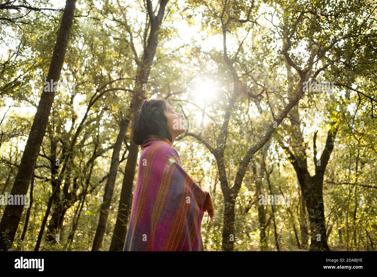 Junge Frau meditiert und macht Yoga im Wald mit Sonnenlicht durch Bäume. Geschlossene Augen. Friedliche Outdoor-Szene. Konzept der Achtsamkeit und Entspannung Stockfoto