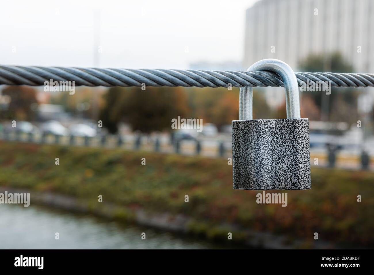 Graues Vorhängeschloss, das am Stahlkabel des Brückenzauns hängt. Unfokussiertes Stadtbild in Herbstfarben im Hintergrund. Horizontale Ausrichtung. Selektiver Fokus. Kopieren Stockfoto