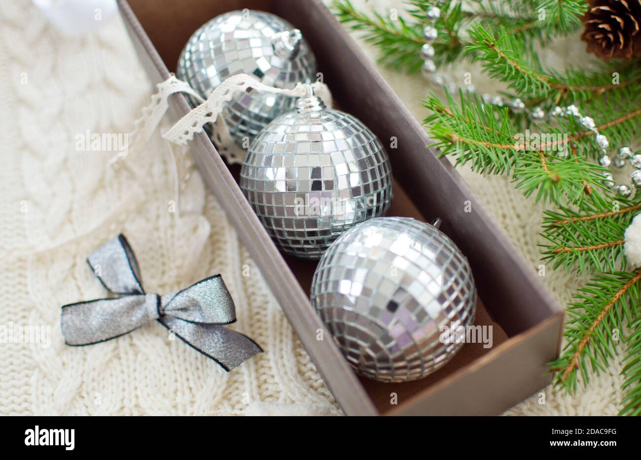 Box mit drei Spiegelkugeln, silbernem Bogen, Gerland, Bändern für Weihnachtsbaumschmuck und Tannenzweig auf weißem gestricktem Hintergrund Stockfoto