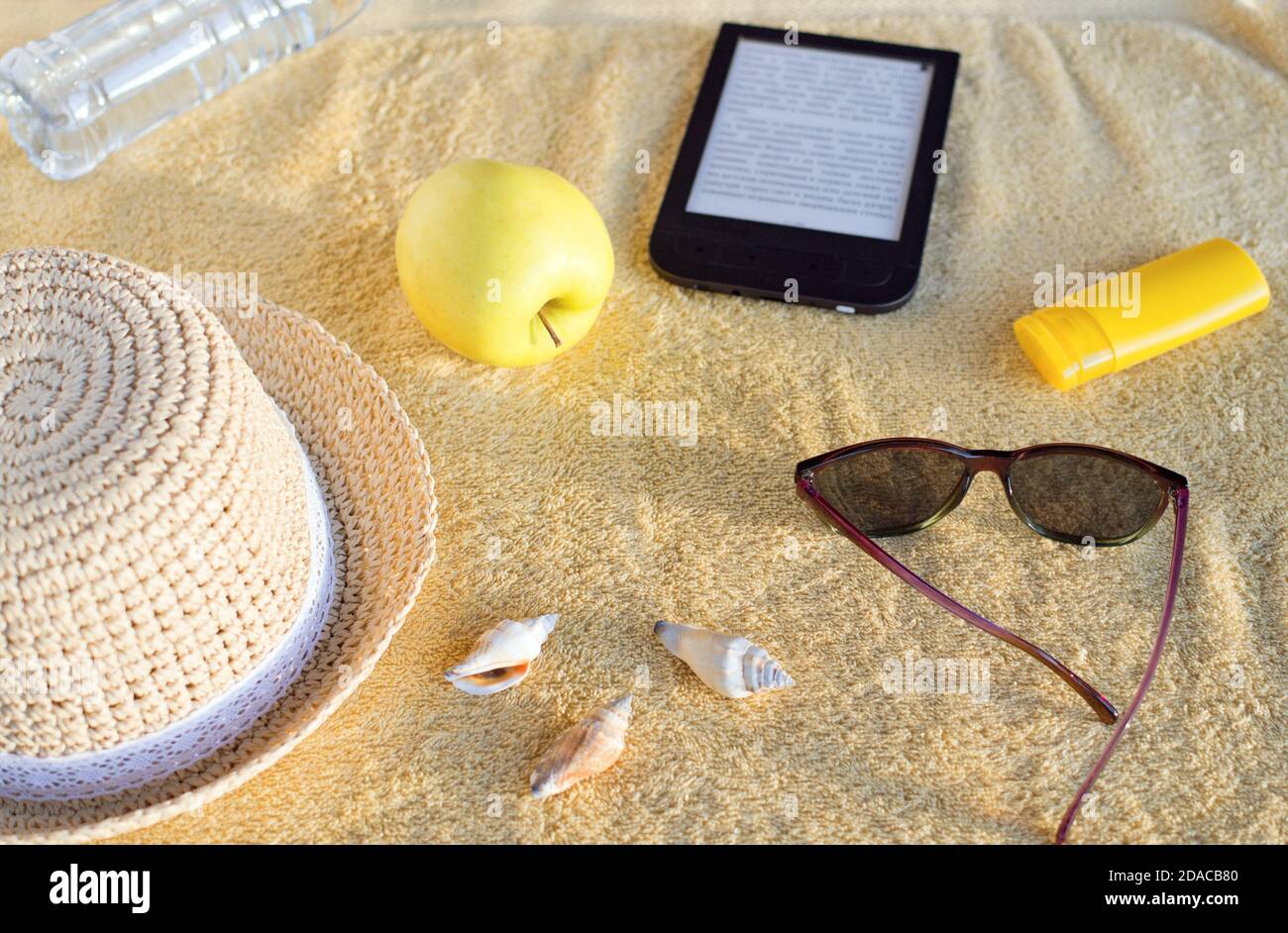 Strohhut, Muscheln, Sonnenbrandcreme, Sonnenbrille, Apfel, Flasche mit Wasser und E-Book auf beigefarbenem Strandtuch. Weiches Sonnenlicht am Abend. Stockfoto