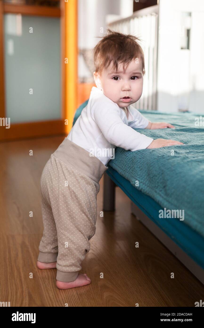 Kleines kaukasisches Kleinkind beginnt zu stehen und zu Fuß auf seinen Beinen, hält sich an der Sofakante Stockfoto