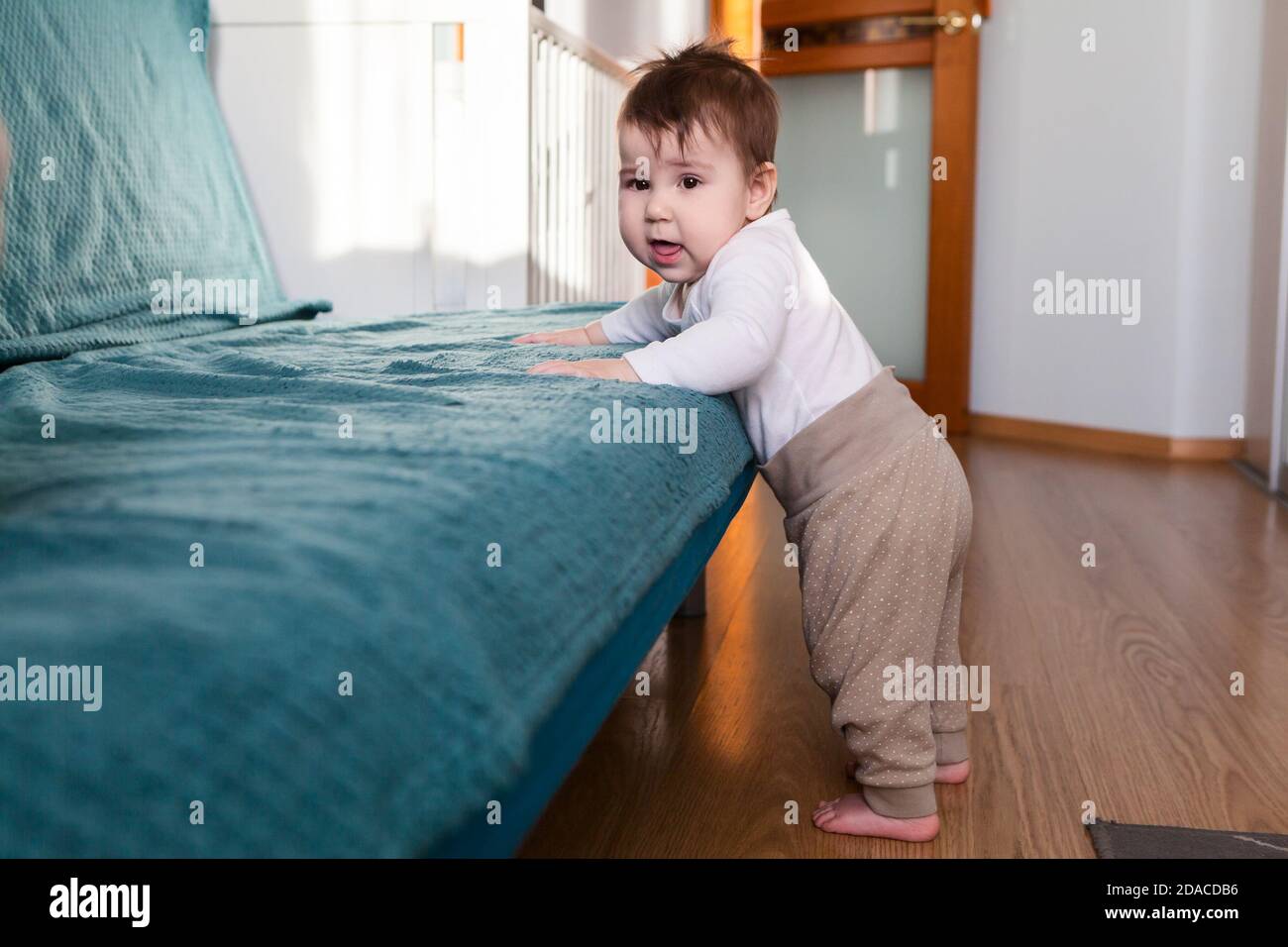 Sieben Monate altes kaukasisches Baby, das auf dem Boden steht, die Hände hält und die Kamera anschaut, das Haushaltszimmer Stockfoto