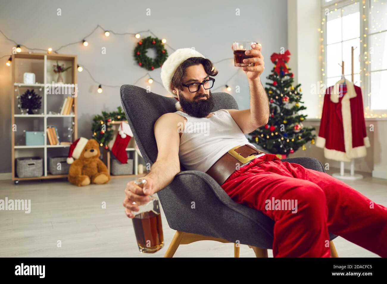 Mann in Santa Mütze und lustige Feiertagskostüm sitzen und Feiern mit Alkohol in Glas und Flasche Stockfoto