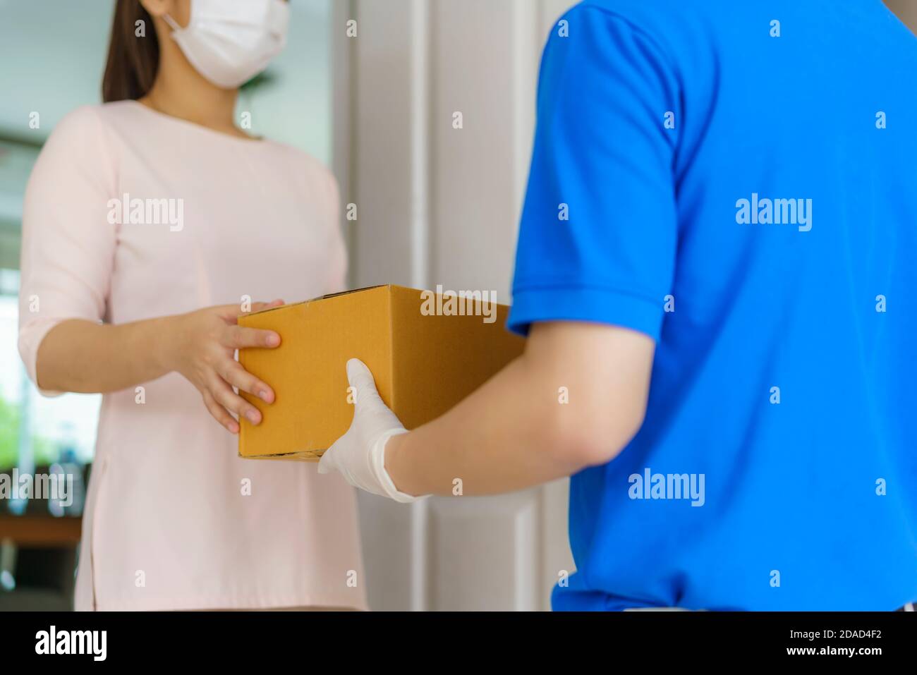 Asiatische Lieferung Mann trägt Gesichtsmaske und Handschuh in blauer Uniform hält einen Karton-Boxen im Vorderhaus und Frau eine Lieferung von Kisten von d Stockfoto