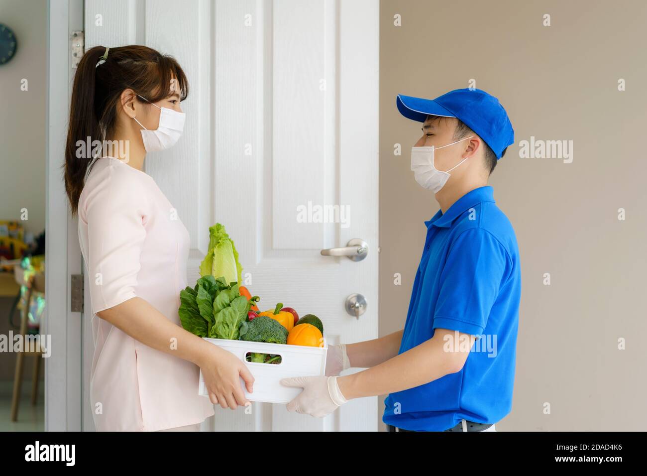 Asiatische Frau Kunde trägt Gesichtsmaske und Handschuh erhalten Lebensmittel-Box mit Lebensmitteln, Obst, Gemüse und trinken von Lieferung Mann vor dem Haus d Stockfoto