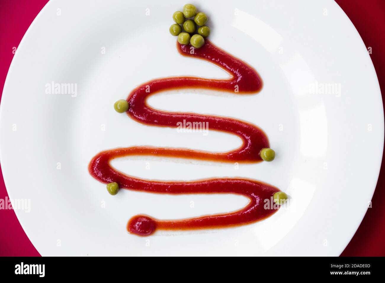 Ketchup auf einem weißen Teller sieht aus wie ein Weihnachtsbaum. Grüne Erbsen dienen als Dekoration. Die Platte steht auf einem roten Hintergrund. Blick von oben Stockfoto