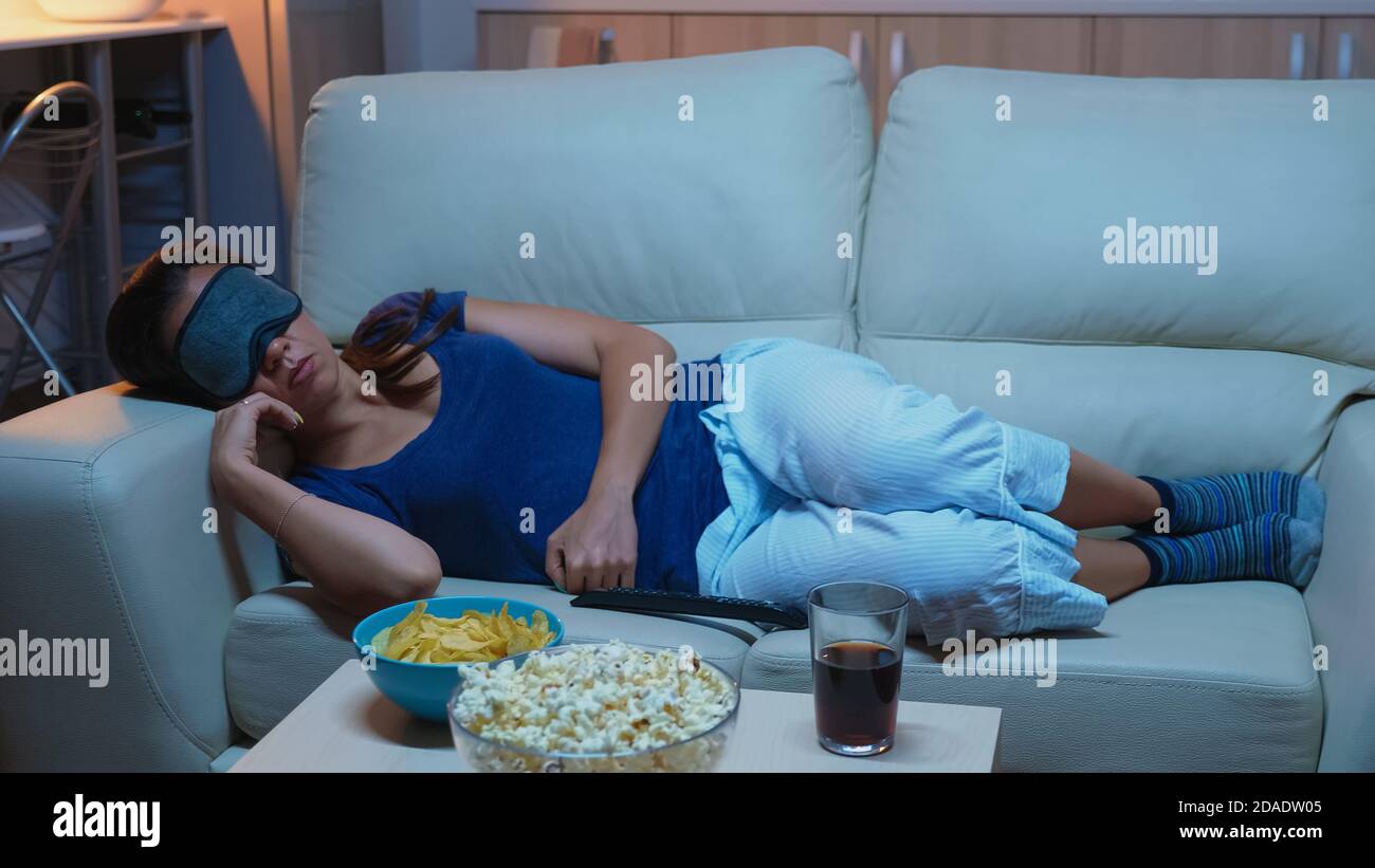Junge Dame, die auf dem Sofa ruht und eine Maske zum Schlafen trägt, liegt auf dem gemütlichen Sofa. Müde erschöpft einsam schläfrig Frau im Schlafanzug Einschlafen auf der Couch vor dem Fernseher, Augen schließen, während Film zu sehen Stockfoto