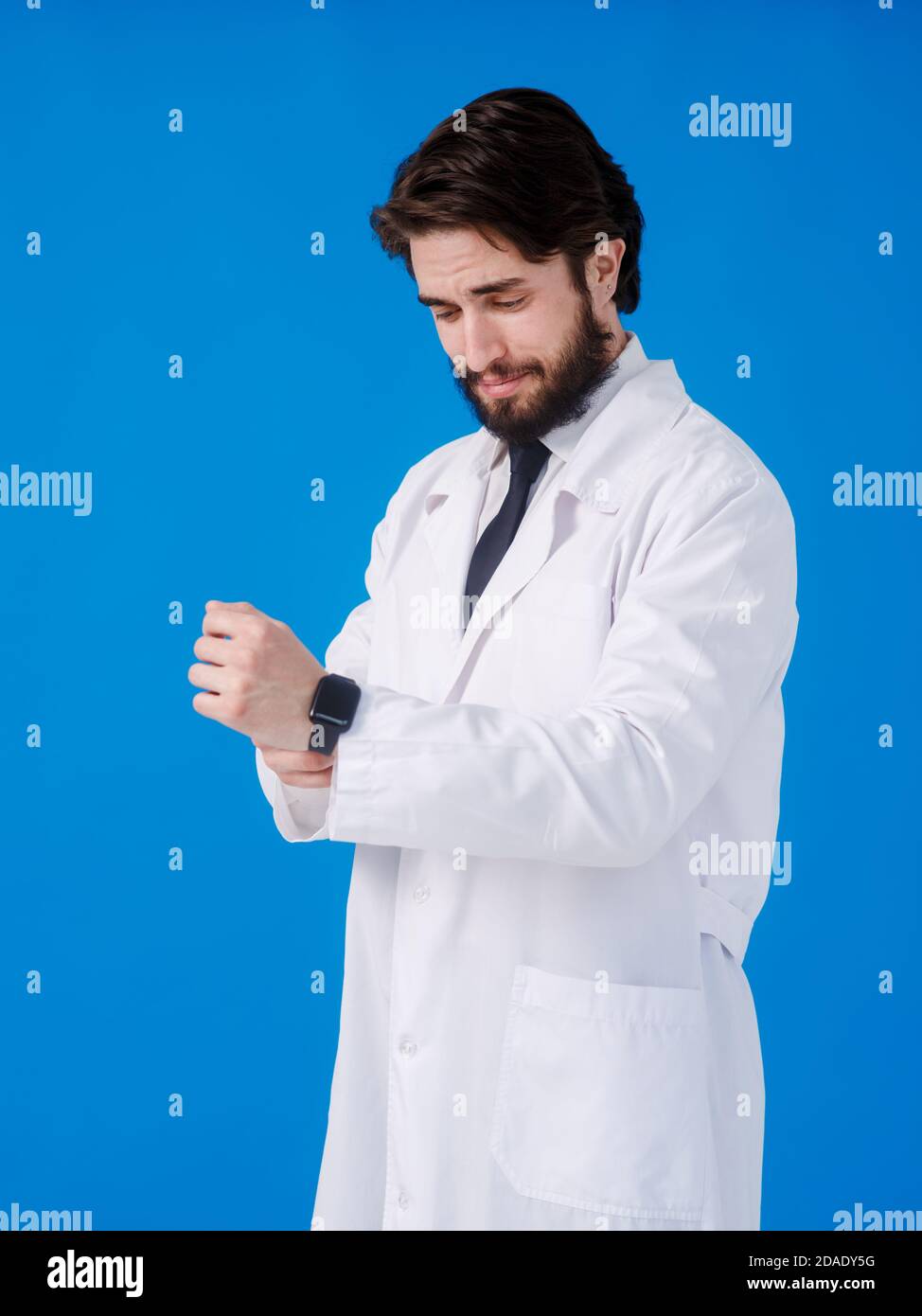 Schöner Arzt Mann mit Bart tragen und Stethoskop über blauem Hintergrund Überprüfung der Zeit auf Armbanduhr, entspannt und zuversichtlich Stockfoto