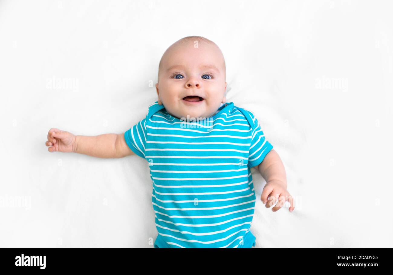 Das positive Kind mit einem blau gestreiften T-Shirt liegt auf einem weißen Bett, lächelt und schaut auf die Kamera. Frohe Kindheit. Draufsicht Stockfoto